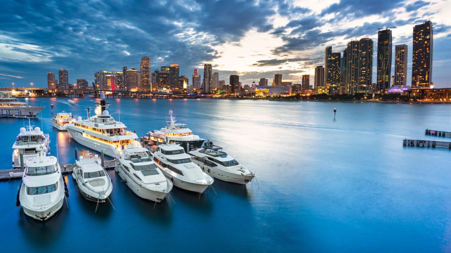 Luxury yachts docked at Biscayne Bay in Miami with the city skyline illuminated at twilight.