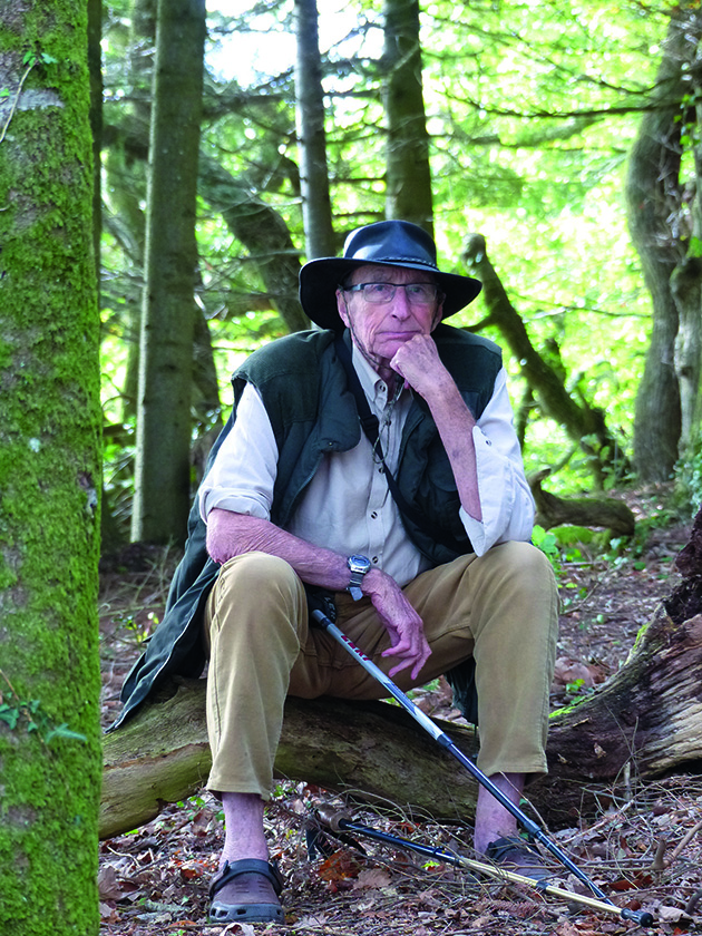 Boat designer and builder James Wharram sitting on a log in woodland